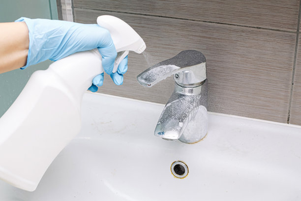 用手将清洁剂喷洒在水槽水龙头上。浴室卫生和消毒过程。戴蓝色手套的人拿着白色瓶子。图片下载