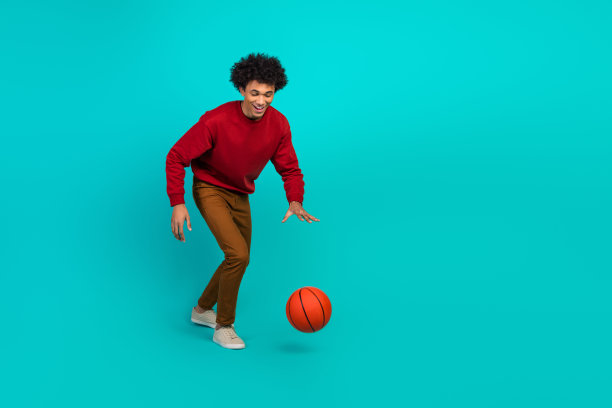 身穿红色毛衣和棕色裤子的年轻男子在绿松石色工作室里运送篮球，拍摄动感的时尚生活方式照片图片下载