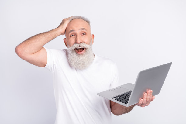 照片：一位快乐、积极、惊讶的老人，一只手扶着头，一手拿着电脑，在灰色背景上感到震惊，孤立。图片下载