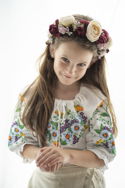 竖直照片，一位戴着花环、身穿白色绣花衬衫的乌克兰女孩，面带微笑，从眉下俏皮地瞥视，手中藏着东西，背景为白色，概念为童年玩耍和乌克兰传统。图片下载