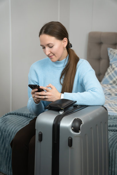 一位年轻的女游客坐在酒店房间的行李箱上，用智能手机查看旅行应用程序，为即将到来的激动人心的旅行和冒险做最后的计划。竖版照片图片下载