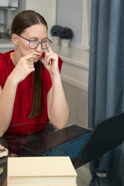 身穿红色衬衫的女性专注于笔记本电脑，正在分析数据。她坐在一张玻璃桌旁，周围摆放着书籍，身处一个明亮、现代的房间里，表情若有所思。竖版照片图片下载