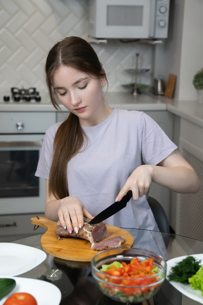 年轻女孩在她现代化的厨房里，在一块木质砧板上小心地切着调味好的烤牛肉，准备一顿健康的餐食，搭配新鲜蔬菜。竖版照片图片下载
