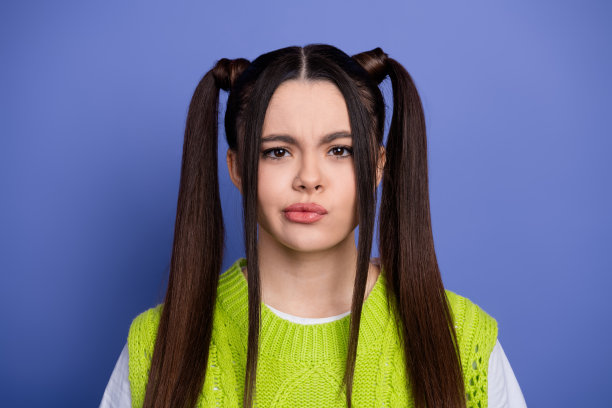 年轻女子扎着马尾辫，穿着亮绿色毛衣，在蓝色背景前微笑并向上指，适合时尚生活方式的图库照片图片下载