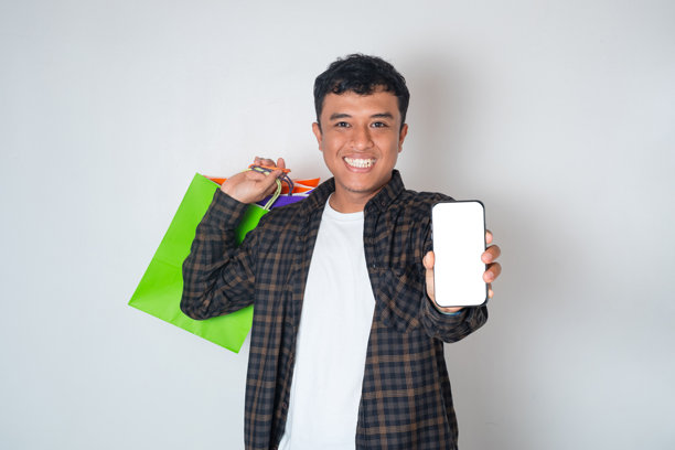 帅气的年轻亚洲男性，手持彩色购物袋，通过手机进行在线购物，手机屏幕为白色空白样机，背景为纯白色。图片下载