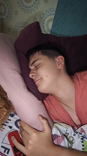 一个少年在床上安详地睡着，手里拿着遥控器，盖着色彩缤纷的毯子。这张图片代表了在家休息、舒适和放松。图片下载