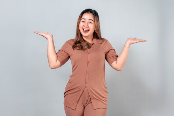 一位快乐的年轻亚洲女性，脸上带着灿烂的笑容，双手高举，做出开朗的耸肩动作。她富有表现力的面孔和愉悦的活力使这张图片成为广告和营销的理想选择。图片下载