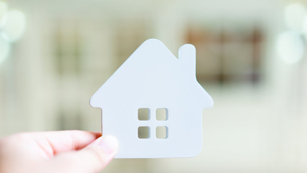 手持白色房屋图标，背景明亮模糊，象征房地产、住房所有权、抵押贷款、住房保险、物业投资和住宅生活方式概念。图片下载