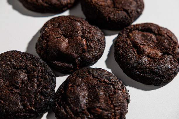 一张详细的特写照片，展示了放在白色表面上的六块黑巧克力饼干，突出其丰富、软糯的质地和深邃的可可色。饼干看起来致密且略带光泽，暗示着有嚼劲的中心。图片下载