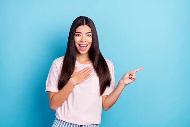 照片：年轻兴奋的女孩，带着快乐积极的笑容，指着蓝色背景上的空白空间图片下载