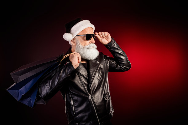 身穿皮夹克、戴着墨镜的摇滚风圣诞老人，提着购物袋，在红灯下摆姿势，为节日圣诞节拍摄的素材照片图片下载