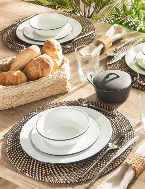 一张乡村户外用餐场景的细节照片，特色是黑色编织餐垫、带黑色边缘的白色搪瓷餐盘和碗、餐具以及一篮新鲜面包。图片下载