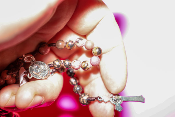 手持宗教念珠手链的特写，念珠由天然粉色和棕色石头珠子、金属奖章和小十字架组成。图片在浅色背景下强调祈祷、灵性和信仰的主题。图片下载