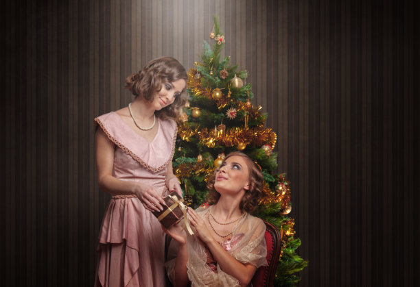 两位身着20年代复古连衣裙的女士在装饰精美的圣诞树前交换圣诞礼物，背景为复古风格。图片下载