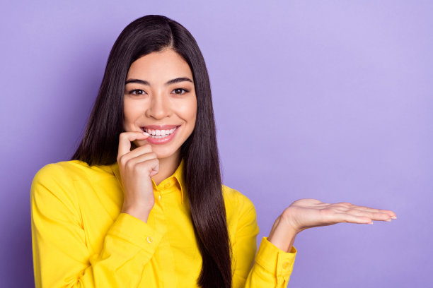 照片：一位留着棕色发型的年轻甜美女士，身穿黄色衬衫，手持空白空间，孤立在紫色背景上，表情调皮图片下载