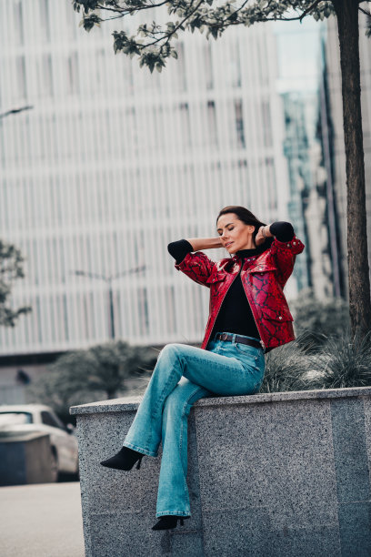穿着红色夹克和休闲牛仔裤的时尚女性在户外摆姿势，在现代建筑旁享受阳光明媚的城市景象，竖幅照片图片下载
