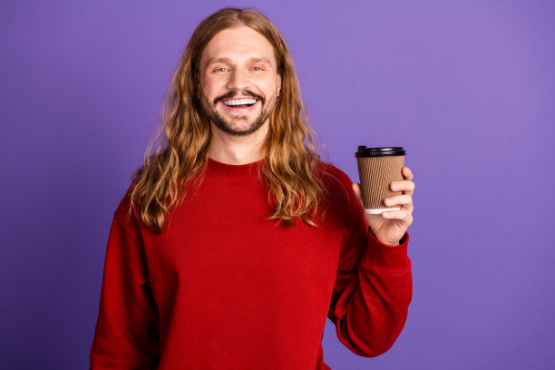 穿着红色毛衣的年轻金发男子，面带微笑，手持咖啡杯，背景为紫色，这是一张休闲时尚生活方式的素材图。图片下载