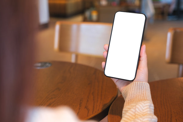 咖啡馆里，一名女子手持并使用着手机，背景是空白的桌面屏幕的样机图。图片下载