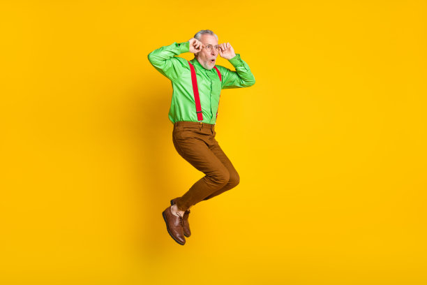 一位令人印象深刻的老年男子全身侧面照片，他戴着眼镜，穿着衬衫和裤子，孤立在黄色的背景上，他跳起来，目光望向空白处。图片下载