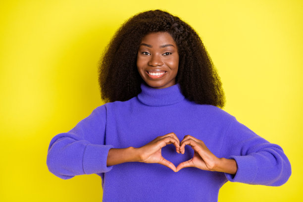 一位女士的手指摆成心形，她穿着紫色的套头衫，背景是黄色的，照片风格。图片下载