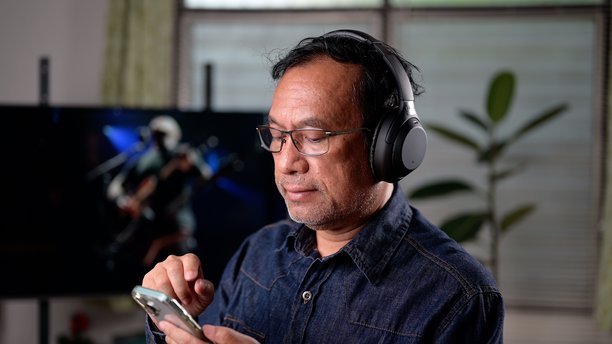 成熟的亚洲男性戴着无线耳机，用智能手机听音乐，在室内放松。休闲生活方式的概念展示了个人享受流媒体、电视。娱乐技术。图片下载