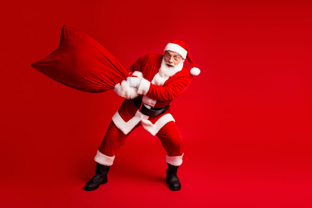 身穿红色圣诞老人服装、背着大麻袋，在亮红色背景前，非常适合圣诞节素材和节日营销活动。图片下载