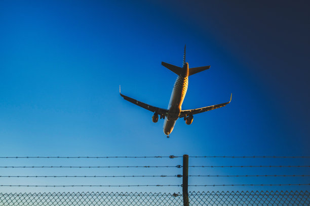 这张图片展示了一架飞机在着陆或起飞过程中，从低空飞越机场围栏的视角拍摄。围栏很可能是机场的安全边界。天空是晴朗的蓝色，表明天气适合飞行。图片下载