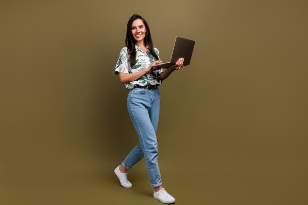 一位快乐友善的人的全身侧面照片，他走在空旷的空间里，手持笔记本电脑，背景是棕色的。图片下载