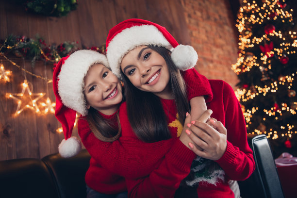 照片：快乐漂亮的两个小姐妹穿着带有红色丑陋印花的新年套头衫，在公寓房间内拥抱在一起。图片下载