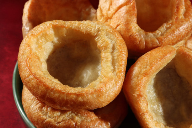 一张全画幅图片，展示了绿色盘子里的酥脆金黄的约克郡布丁（也称泡芙），是搭配丰盛的周日烤肉晚餐的配菜，背景为红色的斑驳图案，焦点在前，俯视角度。图片下载