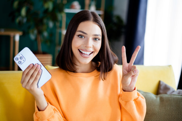 快乐的金发女子穿着橙色毛衣，在舒适的客厅沙发上摆姿势，她拿着手机，做出胜利手势，适合生活方式和房地产图库照片。图片下载