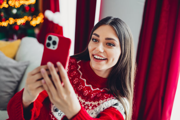 Playful Christmas selfie with a smiling woman in a red knit sweater in a cozy living room beside a festive tree with warm lights图片下载