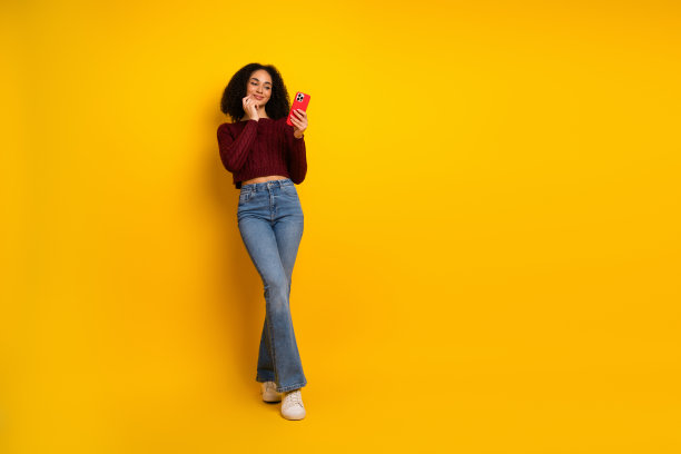 年轻混血女子身穿酒红色毛衣和牛仔裤，对着红色手机微笑，背景是鲜艳的黄色，适合生活方式时尚图库照片图片下载