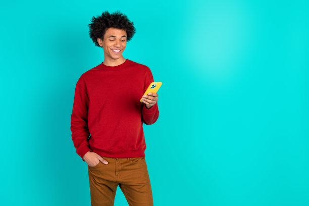 身穿红色毛衣的年轻男子，面带微笑，看着黄色手机，背景为蓝绿色，休闲生活方式素材图图片下载