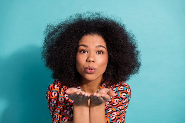 年轻女子留着大大的非洲发型，对着镜头送飞吻，身穿色彩鲜艳的图案衬衫，背景是明亮的蓝色。图片下载