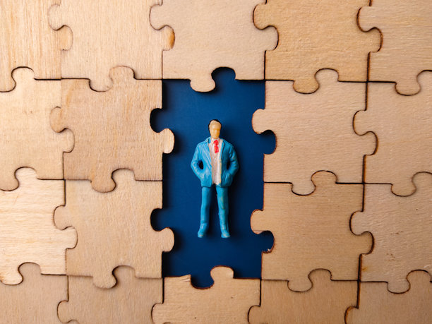 图片描绘了一个商人微缩模型，他站在一个拼图形状的洞里，周围散落着已完成的木制拼图块。图片下载