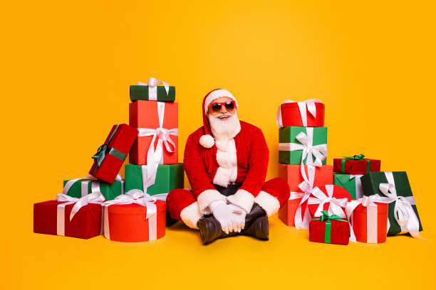圣诞老人穿着红色套装，戴着太阳镜，坐在五颜六色的礼物盒中微笑，为圣诞节拍摄照片图片下载
