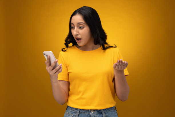 穿着黄色T恤的年轻女子在黄色背景上使用手机的素材照片图片下载