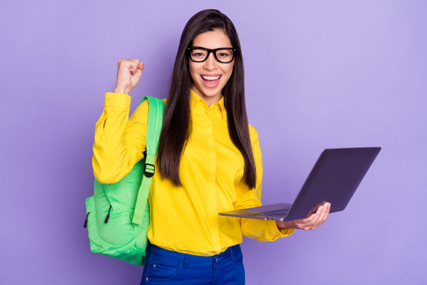 照片：一名快乐、胜利、精神饱满的年轻女子举起拳头，手持笔记本电脑，背景为紫色，展现出获胜者的好心情。图片下载