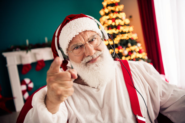 圣诞老人戴着耳机指向镜头，背景是节日客厅，有圣诞树、袜子、花环和礼物，适合作为摄影素材的节日场景图片下载