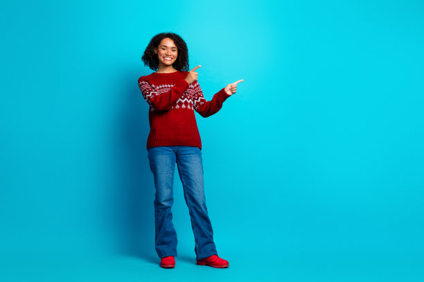 一位年轻的多民族女性穿着红色毛衣、牛仔裤和红色鞋子，在蓝色的工作室背景前摆出姿势，用双手指向两边，照片滑稽而友好。图片下载