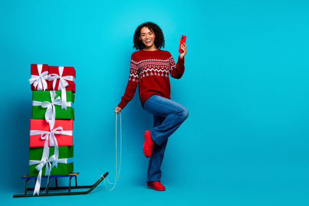 身穿红色毛衣的快乐购物者，驾着雪橇，手捧礼物，在明亮的蓝色背景下自拍，为节日圣诞主题的素材图片。图片下载
