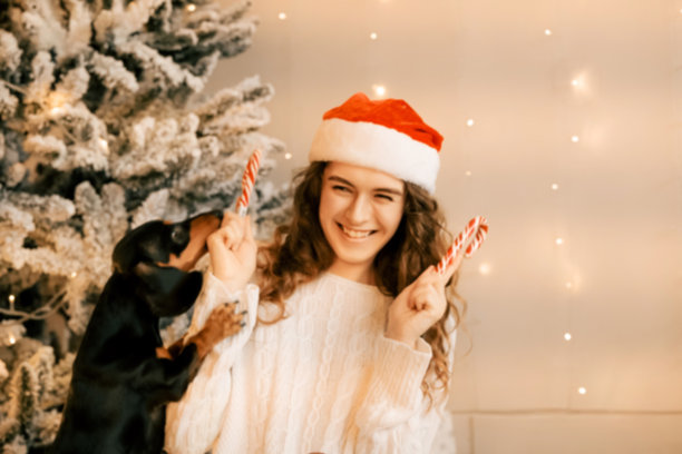 一个美丽的卷发女孩坐在圣诞树旁，怀里抱着一只小狗，这是带有棒棒糖的新年照片拍摄。图片下载