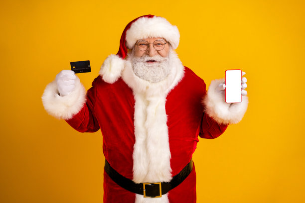 圣诞老人手持信用卡和智能手机，在明亮的圣诞主题照片中，于黄色背景上推广节日购物图片下载