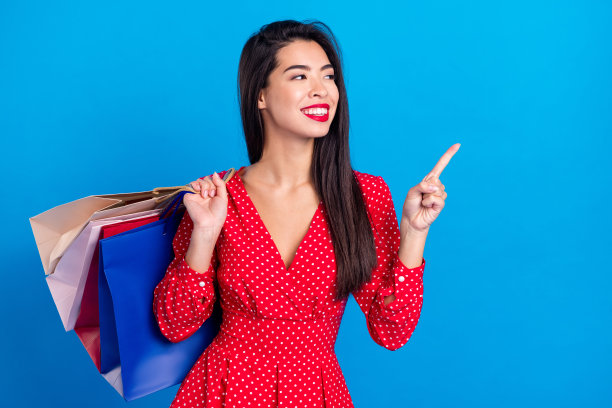 在购物中心展示购物包裹的女性照片肖像，手指指向空白处，背景为明亮的蓝色图片下载