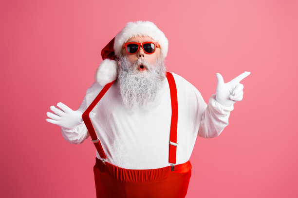 身穿红色吊带和太阳镜的圣诞老人身着粉色工作室，以俏皮的姿势庆祝圣诞节，供节日素材使用图片下载