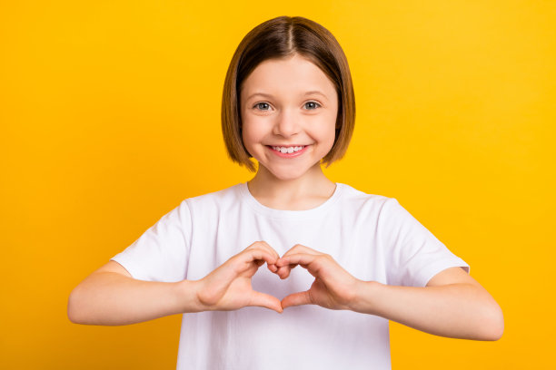 照片肖像：留着波波头的可爱小女孩，在鲜艳的黄色背景前，比出心形手势。图片下载