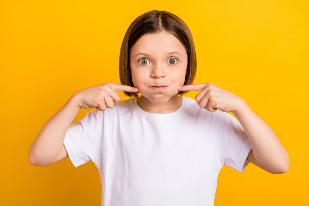 照片肖像：留着波波头的小女孩，屏住呼吸，手指触碰脸颊，俏皮地在明亮的黄色背景前摆姿势图片下载