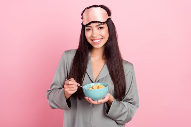 照片：一位迷人的女士戴着睡衣眼罩，微笑着享用装有谷物的盘子和勺子，背景为粉色。图片下载