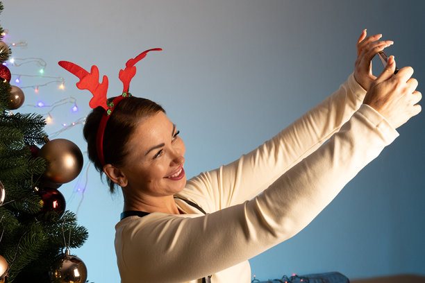 戴着驯鹿角饰的女士正在装饰好的圣诞树旁自拍图片下载